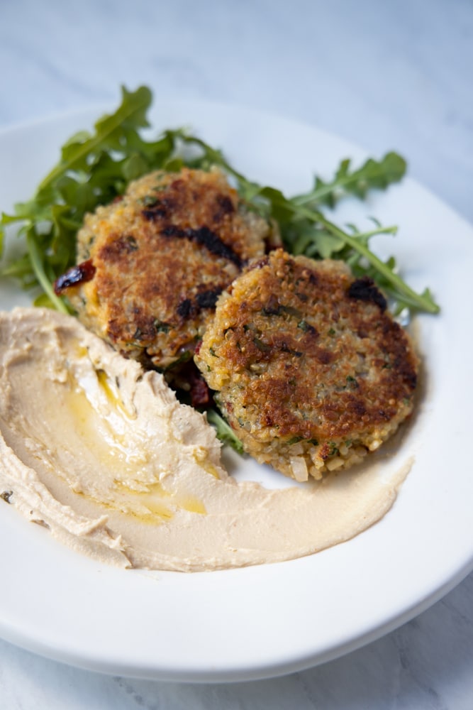 Sundried tomato and basil quinoa patties on a plate with hummus and arugula.