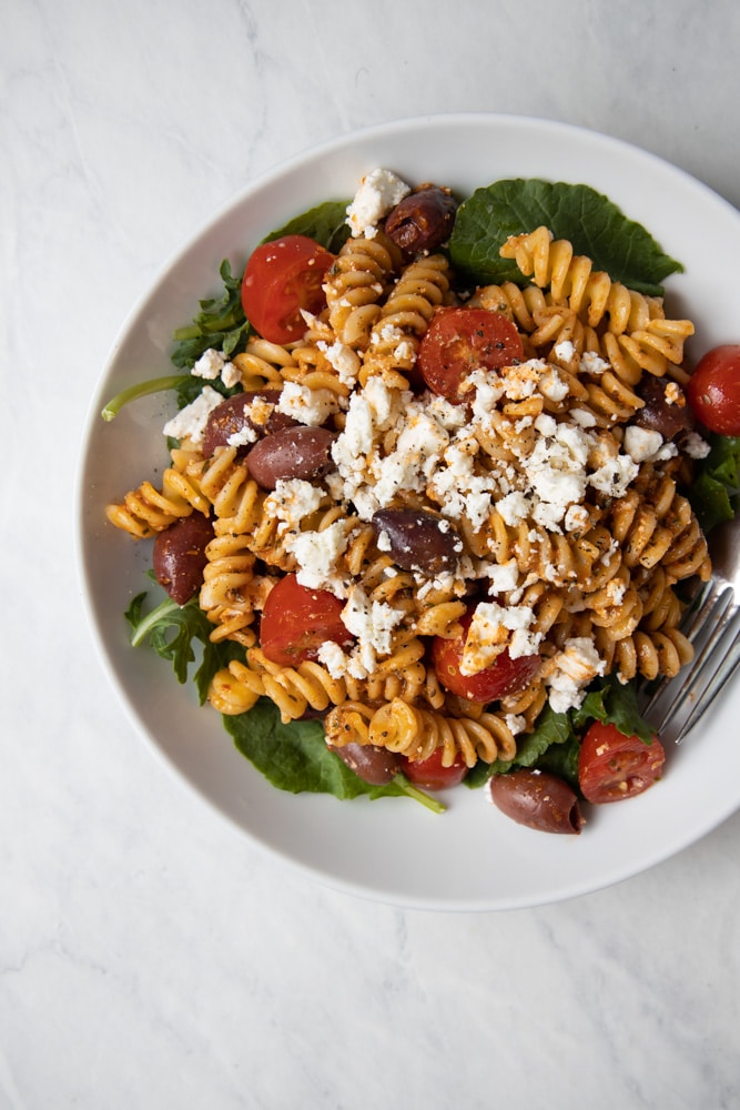 red pepper pasta salad in a bowl with kalamata olives, feta, cherry tomatoes and baby kale