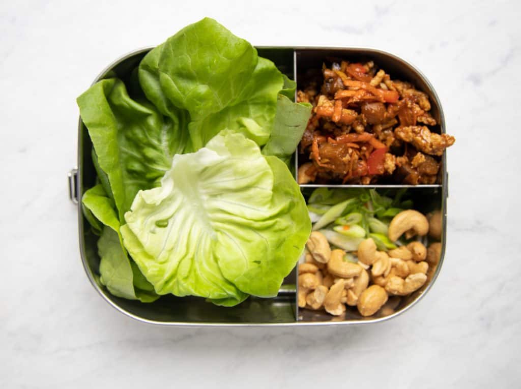 three compartment metal meal prep container with lettuce leaves, chicken mixture and cashews