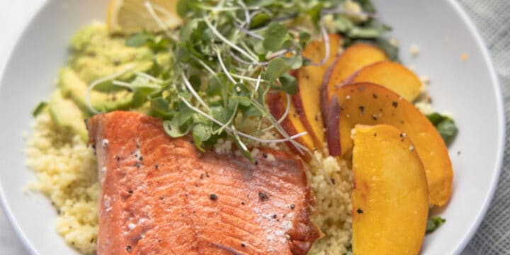 close up image of bowl of quinoa topped with salmon filet, sliced peaches and micro greens