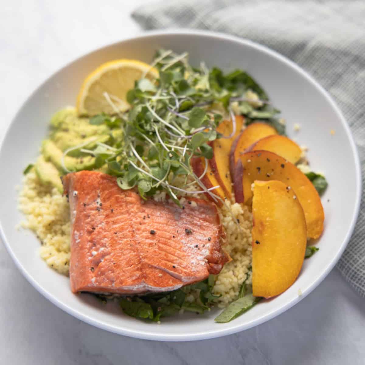 close up image of bowl of quinoa topped with salmon filet, sliced peaches and micro greens