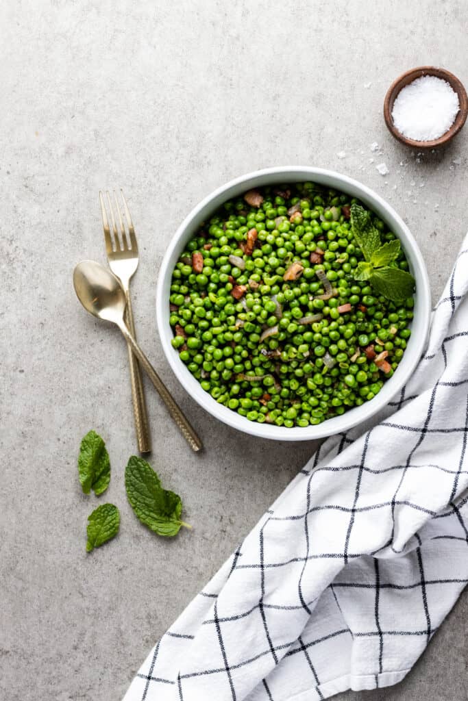 Peas with pancetta in a white bowl with a sprig of mint as a garnish.